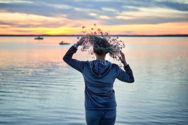 Gölün kıyısında kır çiçeklerinden bir çelenk taşıyan bir kadın. Göl kenarındaki Aziz John arifesinde bir akşam. Litvanya 'da yaz ortası halk festivali