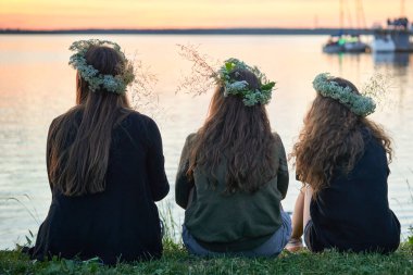 Gölün kenarında kır çiçeklerinden çelenkler taşıyan üç kız. Saint John 's Eve' de göl kenarında bir akşam. Litvanya 'da yaz ortası halk festivali
