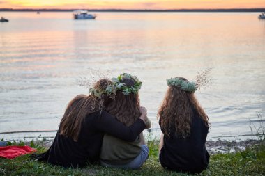 Gölün kenarında kır çiçeklerinden çelenkler taşıyan üç kız. Saint John 's Eve' de göl kenarında bir akşam. Litvanya 'da yaz ortası halk festivali