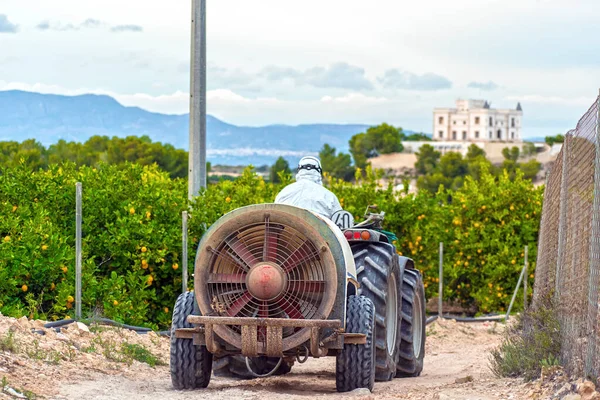 Farmer wearing protective clothes drives a tractor on a lemon plantation in Spain. Farmer about to spray pesticide and insecticide. Weed insecticide fumigation. Organic ecological agriculture. — Stock Photo, Image