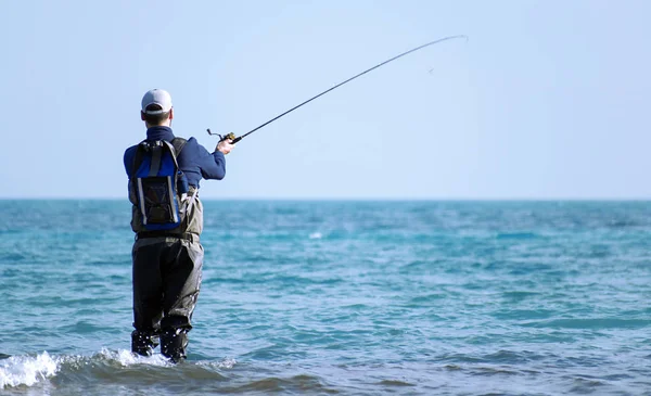 Pescador de pie en la orilla del mar engancha a un pez. Sportsman sostiene una caña de pescar y carrete en un pez enganchado. Spin Pesca hobby la captura de un pez en la playa en Murcia, España, 2019 —  Fotos de Stock