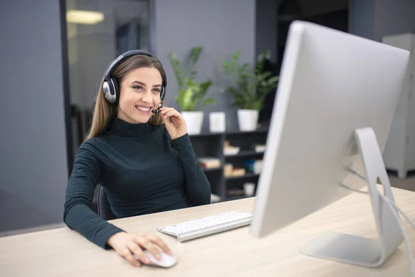 Genç, güzel bir ofis çalışanı internet üzerinden müşteriyle iletişim kurarken gülümsüyor, mikrofonlu kulaklık takıyor.