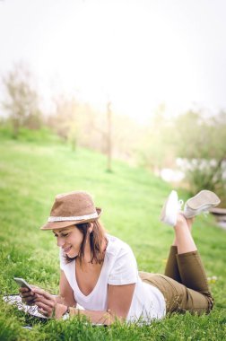 Şapkalı genç bir kadın çimenlerde uzanıyor, dirsekleriyle kendini destekliyor, telefonuna bakıyor. Bacaklar yukarı, çapraz.