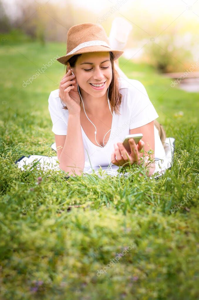 Young happy woman lying in the grass and enjoying music from her phone in the nature. Smiling. Wearing hat.