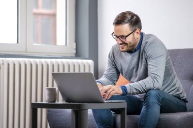 Gözlüklü, genç, yakışıklı, dizüstü bilgisayar kullanan ve evden çalışırken kanepede yan yana oturan mutlu bir serbest yazar. 