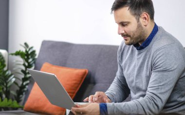 Laptopta çalışan ve evdeki kanepede oturan yakışıklı genç adam.