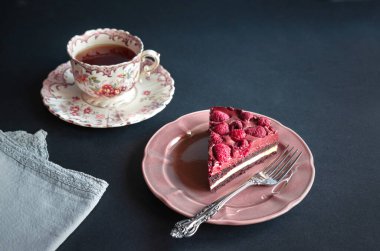 Nefis bir ahududu peynirli kek dilimi güzel bir tabakta ve koyu arkaplanda klasik çiçekli çay fincanı.. 