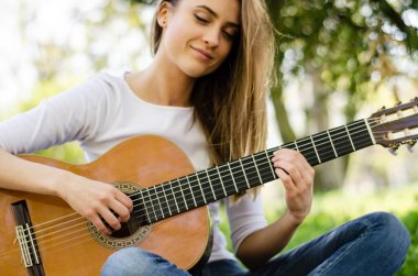 Parkta gitar çalan, yerde oturan sevimli genç kız. Arkasında güneş ışığı.