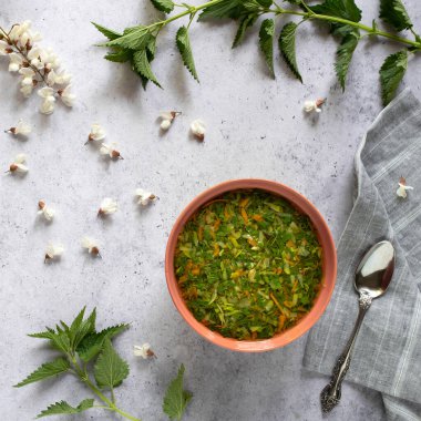 Taze ısırgan otu ve sebzeli bahar çorbası kırsal bir kase ve peçete, yukarıdan manzara. Akasya çiçekleri ve ısırgan otlarıyla süslenmiş. Rustik desenli arkaplan. 