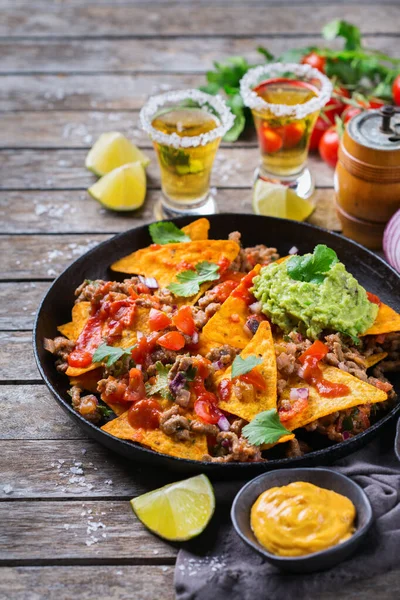 Yellow corn tortilla chips nachos with ground beef, mince, guacamole, red hot jalapeno chili salsa and cheese sauce with tequila on a wooden table.