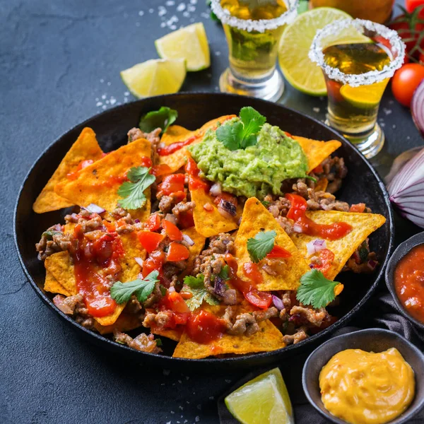 Yellow corn tortilla chips nachos with ground beef, mince, guacamole, red hot jalapeno chili salsa and cheese sauce with tequila on a dark table.