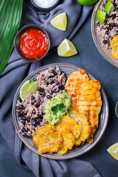 Traditional Central American caribbean cuban colombian food. Rice with black beans, roasted fried chicken breast and tostones, fried green bananas plantains with guacamole sauce. Top view, flat lay