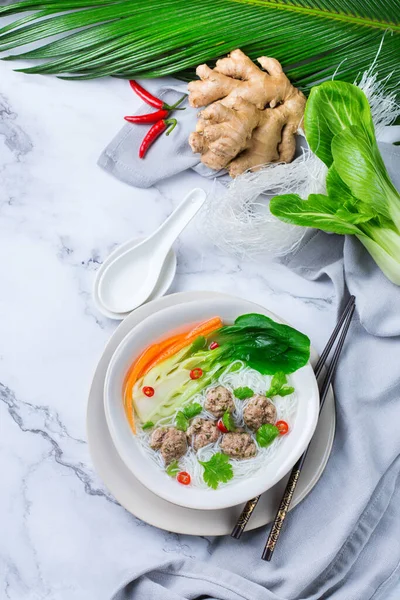 Food and drink, traditional asian cuisine. Spicy bee hoon, bihun soup with pork meat balls, bok choy and rice vermicelli noodles on a marble table. Top view flat lay background — Stock Image Food Drink Traditional Asian Cuisine Spicy Bee Hoon Bihun Soup Stock Photo