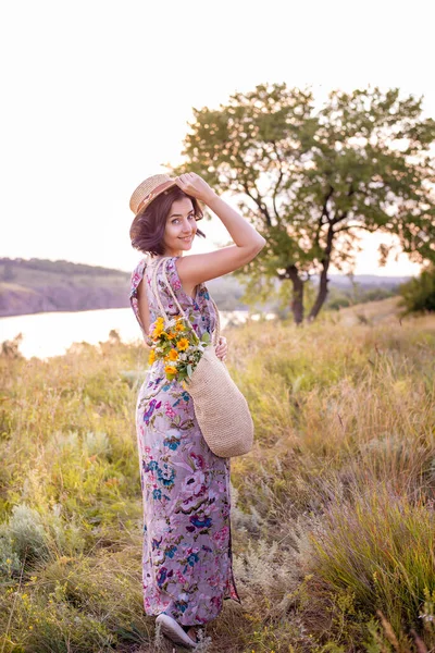 Look from back of romantic short haired brunette woman with flowers in bag and straw hat in hand walking in field
