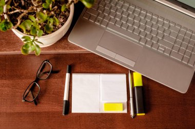 Geçmişteki boş, beyaz notalar ve sarı etiketler, gözlük, kalem, keçeli kalem, keçeli kalem, keçeli kalem ve bitki ile çevrili ahşap masaüstü. Dizüstü bilgisayarının klavyesinde düz bir çalışma alanı. Anlaşıldı. Sabah ofiste..