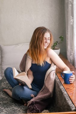 Mutlu, güzel, beyaz, gülümseyen bir kız seksi, çıplak omuzlu bir kanepede dinleniyor, kitap okuyor, elinde bir fincan kahve / çay tutuyor. karantina zamanında sıcak ekoseli kapalı bir yerde. Ev kavramında kalın.