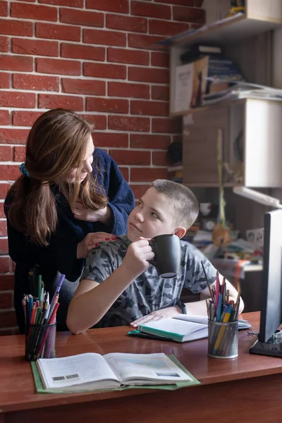 Mother put hands on shoulders of son with a cup of tea as help of doing homework,studying at home.Distance education in quarantine.Good relationship between mom and son indoor.Smiling caucasian family