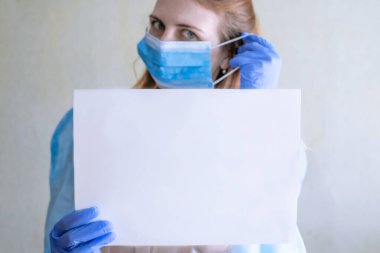 Beyaz tenli güzel bir kız koruyucu yüz maskesi, mavi nitril cerrahi eldiven ve tıbbi önlük giyerek beyaz bir not tutar. Coronavirus koruması. Yazı için yer ayır..