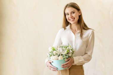 Gülümseyen güzel kız, arka planında beyaz mersin çiçekleri ve makaronlar olan mavi hediye kutusunu tutuyor. İş dünyasından bir kadın doğum gününde parlak ve muhteşem bir buket hediye alıyor.