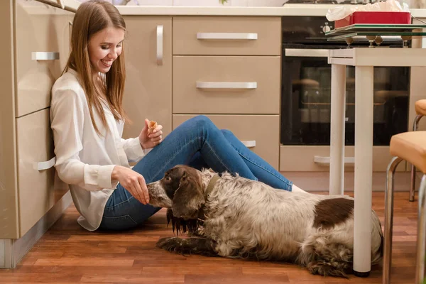 Young smiling woman sit on floor and feed dog from hand in kitchen.Caucasian girl spending playful time with domestic pet friend isolated indoor.Russian spaniel is member of family lying on the floor.