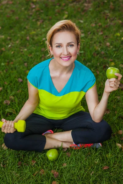 Sportliche Frau mit Hantel und grünem Apfel sitzt auf einer Wiese. — Stockfoto