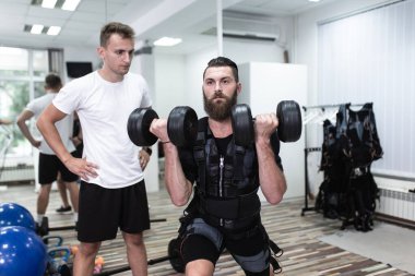 Genç sakallı adam, rehabilitasyon merkezinde kişisel antrenörüyle birlikte elektrik kas stimülasyonu kıyafeti üzerinde egzersiz yapıyor..