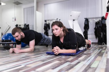 Genç bir kadın, rehabilitasyon merkezinde kişisel antrenörüyle kas stimülasyonu elbisesi üzerinde egzersiz yapıyor..