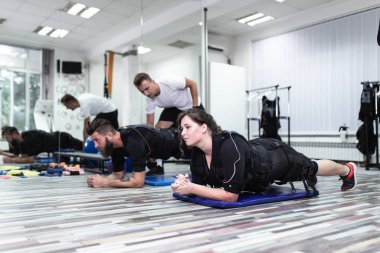 Genç sakallı adam ve kadın, rehabilitasyon merkezinde kişisel antrenörleriyle birlikte elektrik kas stimülasyonu kıyafeti üzerinde egzersiz yapıyorlar..