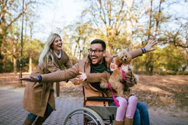 Tekerlekli sandalyedeki engelli baba parkta kızı, karısı ve köpeğiyle eğleniyor..