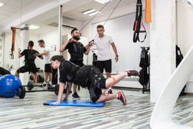 Genç sakallı adam ve kadın, rehabilitasyon merkezinde kişisel antrenörleriyle birlikte elektrik kas stimülasyonu kıyafeti üzerinde egzersiz yapıyorlar..