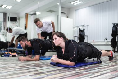 Genç sakallı adam ve kadın, rehabilitasyon merkezinde kişisel antrenörleriyle birlikte elektrik kas stimülasyonu kıyafeti üzerinde egzersiz yapıyorlar..