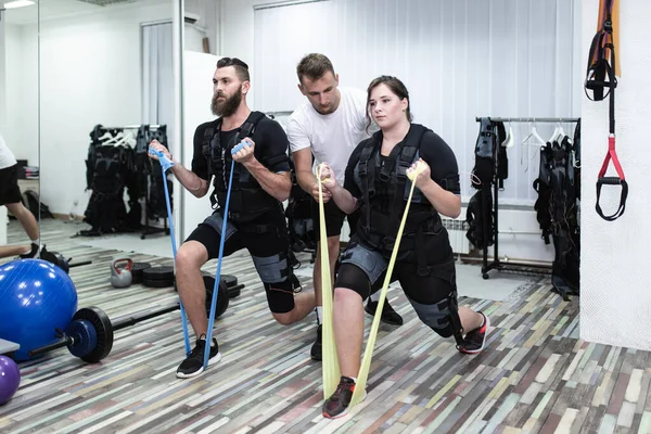 Young bearded man and woman doing exercises in electrical muscular stimulation suit with their personal trainer at rehabilitation center.