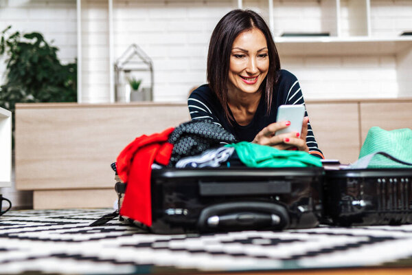 Красивая молодая женщина упаковывает свои вещи в большой чемодан и смотрит на мобильный телефон. Концепция подготовки путешествий
. 