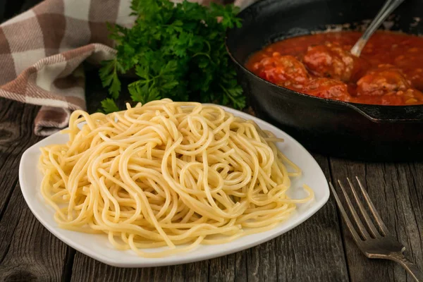 Cierre los espaguetis en un plato y una sartén con albóndigas en salsa de tomate sobre un fondo rústico de madera —  Fotos de Stock