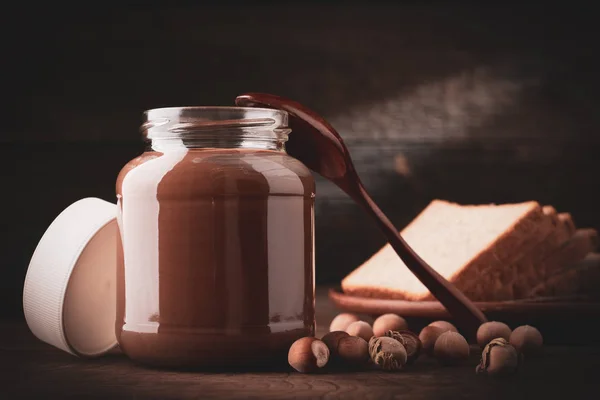 Банка шоколадно-ореховой пасты с ломтиками свежего белого хлеба и фундука на деревянном фоне — стоковое фото
