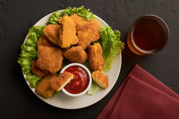 Placa com vista superior de nuggets de frango cozido com ketchup e um copo de cerveja em um fundo escuro — Fotografia de Stock