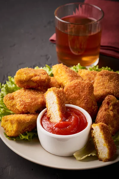 Uma fatia de nuggets de frango em ketchup em um prato cheio de nuggets de frango e um copo de vinho em um fundo escuro tiro vertical — Fotografia de Stock