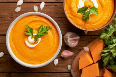 İki kâse balkabağı çorbası ve ahşap arka planda kesme tahtasındaki çiğ balkabağı manzarası.