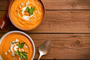 İki kâse kabak çorbasının üst görüntüsü kabak tohumları ve kaşık serpiştirilmiş ahşap bir arka planda