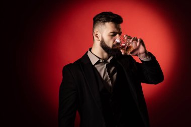 Yakışıklı sakallı, şık sakallı bir adam elinde viski bardağıyla kırmızı arka planda onu içecek.