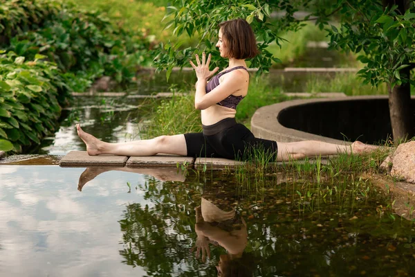 Young woman doing yoga in the morning park, near a small decorative lake. The figure of a woman is reflected in the water. Summer day — Stock Photo, Image