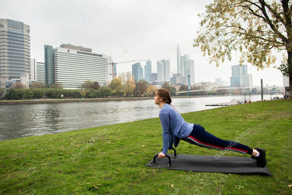 Young athlete woman doing a push up with push-up bars on the grass, near the river, on the background of the city. Sport and training concepts.