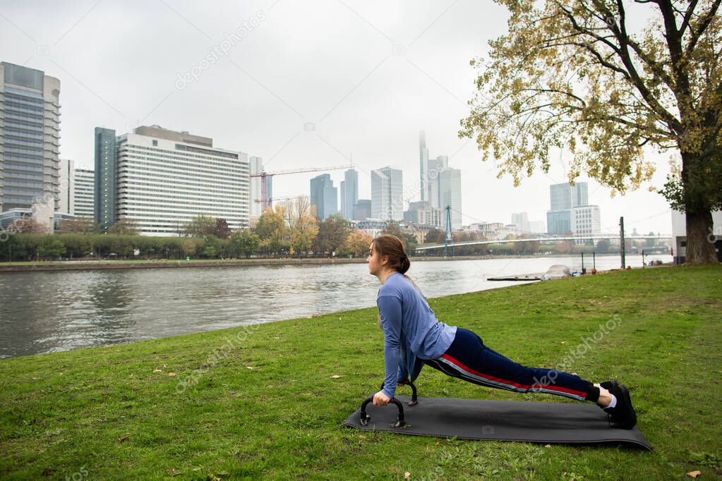 Young athlete woman doing a push up with push-up bars on the grass, near the river, on the background of the city. Sport and training concepts.