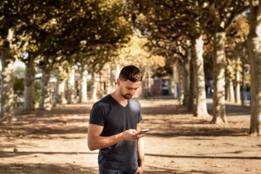 Tişörtlü genç adam elinde cep telefonuyla sokakta duruyor ve mesaj yazıyor. Arka planda bir ağaç şeridi var. Frankfurt, Almanya.