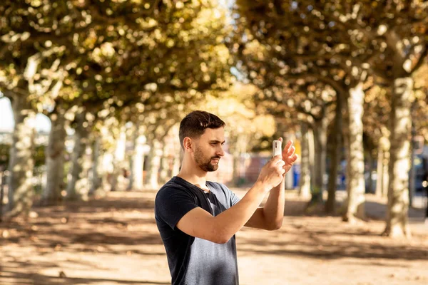 穿着T恤的年轻人站在街上 用手机拍照 后面是一条林荫道 德国法兰克福 — 图库照片
