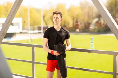 Stadyumun yanında spor yaptıktan sonra havluyla gezen sağlıklı genç bir adam. Aktif yaşam tarzı, spor konsepti.