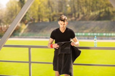 Stadyumun yanında spor yaptıktan sonra havluyla gezen sağlıklı genç bir adam. Aktif yaşam tarzı, spor konsepti.