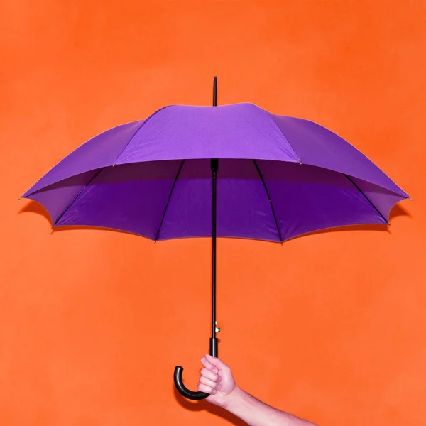 Ein Mann hält in der Hand einen lila Regenschirm auf dem Hintergrund einer orangefarbenen Wand. Konzept Herbst, Wirtschaft, Kunst. — Stockfoto