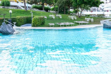 Yanı başında turkuaz su bulunan bir havuzu olan otelin manzarasında binaların arka planında çimenlerin üzerinde güneş panoları var. Yaz konsepti, rahatlama, spa, su parkı, manzara tasarımı.