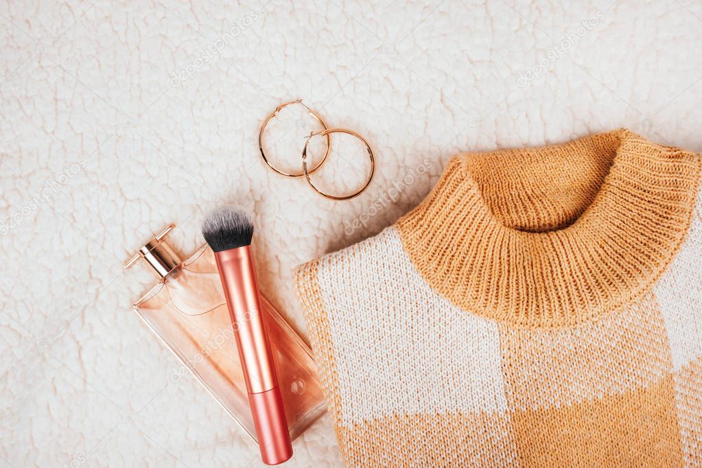 Fashion flatlay with beige sweater, perfume bottle, make-up brush and trendy earrings on cozy off white blanket.